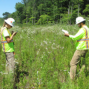 two GEI employees with hard hats and yellow vests monitoring in a field